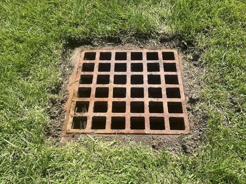 Close-up of a metal storm drain inlet grate set in a grassy area, showing a cleared opening and surrounding turf after maintenance and cleaning work.